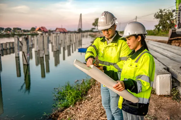 İki erkek ve kadın mühendis nehirdeki kazıkların inşaat planlarını ve ilerlemesini inceliyor..