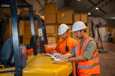 Bir depoda forkliftin arkasında durup kutuları kontrol eden ve not alan iki paket müfettişi var..