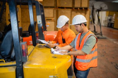 İki depo müfettişi kutulara bakmak ve not almak için bir forkliftin arkasında duruyorlar..