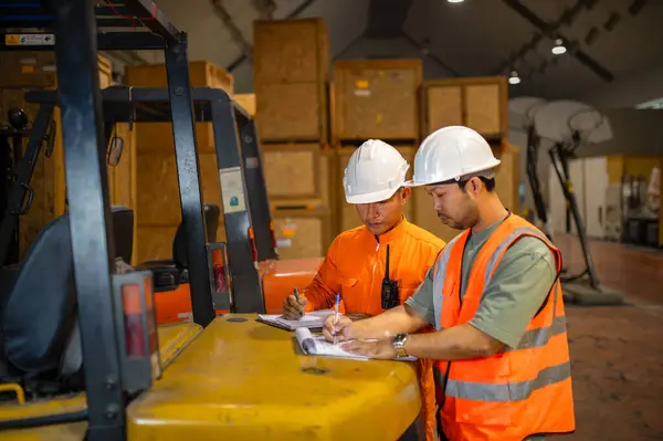 Bir depoda forkliftin arkasında durup kutuları kontrol eden ve not alan iki paket müfettişi var..
