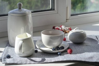 Beyaz kupa ve penceresinin arkasında böğürtlen olan tatlı. Güneşli bir sabahta böğürtlenli kahvaltı, tatlı olarak yaban mersini ve ahududu. Kahvaltı için hazırlanmış beyaz tabaklar.