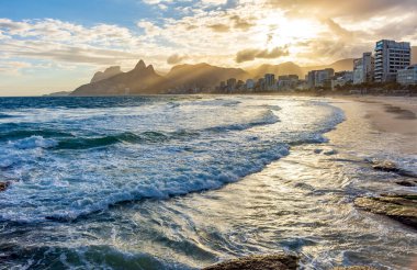 Rio de Janeiro 'daki Ipanema plajında gün batımı. Önünde binaları, arkasında dağları var.