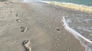 Sakin bir sahil boyunca yumuşak deniz dalgalarıyla kumdaki ayak izleri. Sıcak ve sakin bir günde, yumuşak köpükle kıyıya değen huzurlu bir sahil anı.