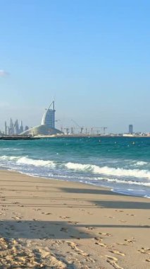 Dubai 'de güzel bir sahil manzarası, dalgalar kıyıya vuruyor, açık mavi deniz, ve Dubai ikonu Burj Al Arab ve Dubai Marina gökyüzü altında görülebiliyor.