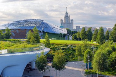 Zaryadye Park, Moskova 'da sakin bir sabah, ön planda yemyeşil bir yer ve yumuşak bir yaz gökyüzünün altında duran Stalinist bir gökdelen.