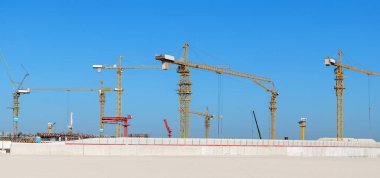 Açık mavi gökyüzünün altındaki büyük bir inşaat alanında birden fazla vinç var. Görüntü, ağır makineler ve yapısal büyüme ile çağdaş inşaat projelerinin ölçeği ve yoğunluğunu vurguluyor