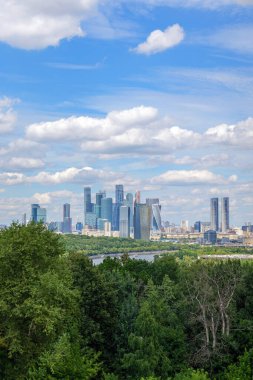 Vorobyovy Gory 'nin Moskova Nehri üzerindeki manzarası, yemyeşil ağaçlar ve mavi gökyüzündeki bulutlar. Moskova Nehri 'nin doğal güzelliği arka planda şehrin ufuk çizgisini tamamlar.
