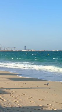 Dubai 'de güzel bir sahil manzarası, dalgalar kıyıya vuruyor, açık mavi deniz, ve Dubai ikonu Burj Al Arab ve Dubai Marina gökyüzü altında görülebiliyor.