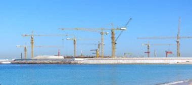 Deniz kıyısındaki büyük bir inşaat sahasında çok sayıda vinç ağır makineler ve su kenarındaki modern yapısal genişleme ile kıyı gelişiminin ölçeğini ve ilerlemesini gösteriyor.