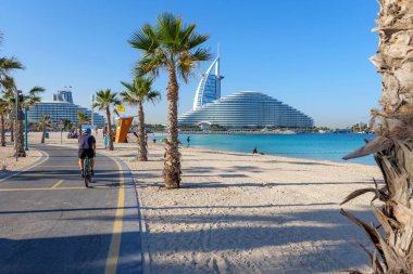 Burj Al Arab 'ın Dubai, BAE' deki Kite Beach boyunca uzanan palmiye çizgili bisiklet yolunun manzarası. İkonik mimari, lüks ve sahil cazibesini birleştiren modern bir şehir manzarası. 24 Ocak 2025, Dubai, BAE