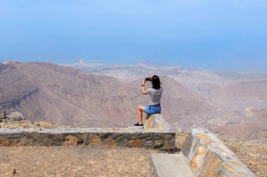 Taştan bir bariyerin üzerinde oturan kadın Birleşik Arap Emirlikleri 'nin en yüksek zirvesi olan Jebel Jais' ten nefes kesici bir dağ ve deniz manzarası çekiyor.