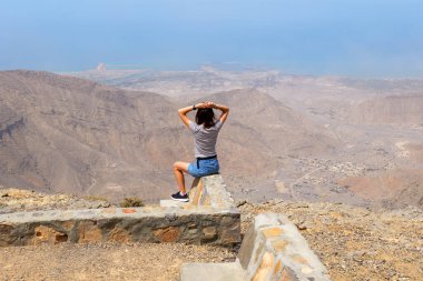Jebel Jais 'in tepesinde taş bir duvarda oturan kadın aşağıdaki geniş dağ manzarasının ve kıyı şeridinin tadını çıkarıyor. Ras Al Khaimah dağlarında manzara, huzur, nefes kesici an, BAE