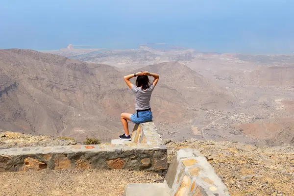 Jebel Jais 'in tepesinde taş bir duvarda oturan kadın aşağıdaki geniş dağ manzarasının ve kıyı şeridinin tadını çıkarıyor. Ras Al Khaimah dağlarında manzara, huzur, nefes kesici an, BAE