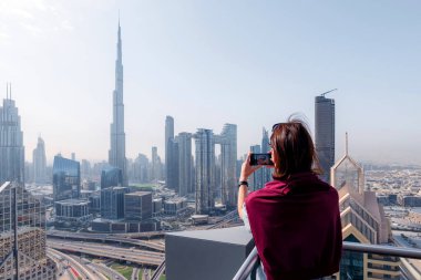 Arka planda Burj Halife ile modern Dubai siluetinin fotoğrafını çeken kadın. Açık bir günde çatıdaki terastan şehir manzarası. Turist manzaranın tadını çıkarıyor. 13 Şubat 2025, Dubai, BAE