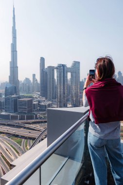 Arka planda Burj Halife ile modern Dubai siluetinin fotoğrafını çeken kadın. Açık bir günde çatıdaki terastan şehir manzarası. Turist manzaranın tadını çıkarıyor. Dikey resim