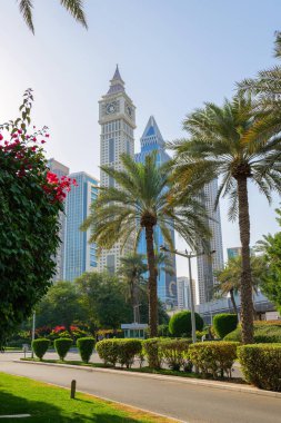 11 Şubat 2025, Dubai, BAE. Önünde palmiye ağaçları ve şehir yeşili olan Emirates Kuleleri ve Saat Kulesi manzarası. Modern gökdelenler, açık mavi gökyüzü ve güneşli hava