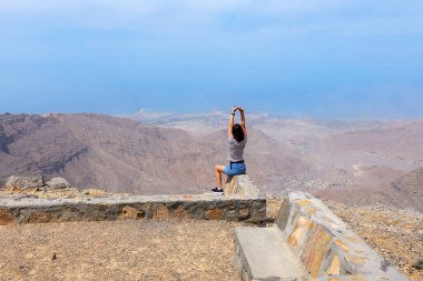 Jebel Jais 'in tepesinde taş bir duvarda oturan kadın aşağıdaki geniş dağ manzarasının ve kıyı şeridinin tadını çıkarıyor. Ras Al Khaimah dağlarında manzara, huzur, nefes kesici an, BAE