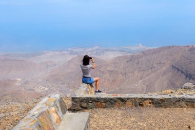 Taştan bir bariyerin üzerinde oturan kadın Birleşik Arap Emirlikleri 'nin en yüksek zirvesi olan Jebel Jais' ten nefes kesici bir dağ ve deniz manzarası çekiyor.