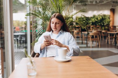 White brunete woman using smartphone while sitting in cafe indoors. Young woman at cafe drinking coffee or tea indoors on a rainy day. Concept of breakfast.