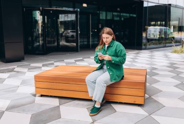 Genç mutlu kadın gülümsüyor ve şehirde akıllı telefon kullanıyor. Güzel bir kadın şehrin caddesinde bankta oturuyor ve akıllı telefonuyla internetten bilgi arıyor..