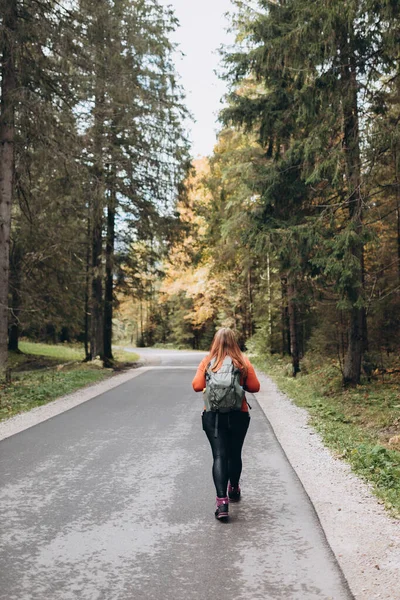Solo tourist Woman hiking on footpath in autumn forest. Full body photo. Happy redhead person in active trekking clothes walking on nature backgraund.