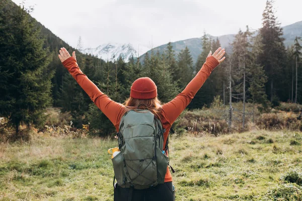 stock image Back view of stylish woman, wearing green backpack and red hat, looking at mountain view while relaxing in nature. Travel and wanderlust concept