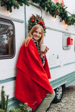 Sarışın kadın kampta bir fincan kahveyle ısınıyor. Modern karavan karavanının yanında duran neşeli genç kız. Noel konsepti.