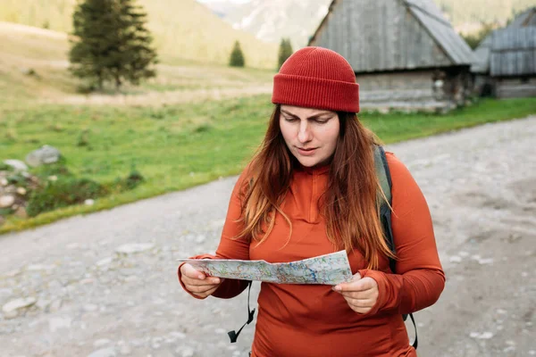 Sad female tourist is exploring new locations on mountains background. Unhappy girl holding a paper map in a sunny day. Travel concept. Hiking woman wearing knit hat exercising outdoor
