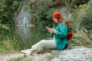 Sırt çantalı şık bir kadın akıllı bir telefon tutuyor ve doğada dinleniyor. Tam vücut fotoğrafı. Aktif kadın yaşam tarzı yürüyüşü. Açık havada oturmak..