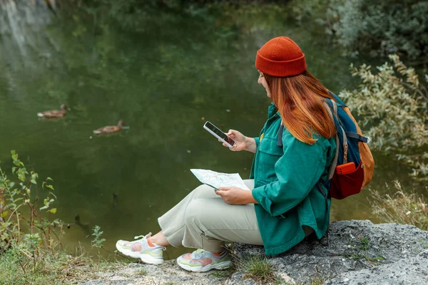 スマートフォンや紙の地図を屋外で保持緑のバックパックを持つスタイリッシュな女性 岩の上に座って活発な女性のライフスタイルをハイキング 赤い帽子をかぶっている女の子自然湖の背景にリラックス — ストック写真