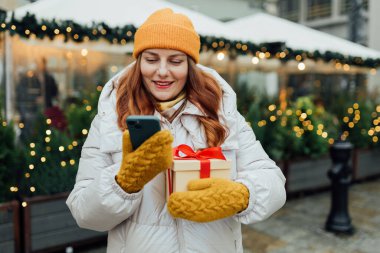 Elinde bir kutu hediye tutan ve Noel süslemeleri yakınlarındaki telefondan mesaj gönderen mutlu genç bir kadın.