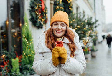 Bir kadın kışın dışarıda bir Noel hediyesi tutuyor. Kar yağışı. Firavun ağacı, çelenk arka planda. Alışveriş, tatil ve Noel kavramı.