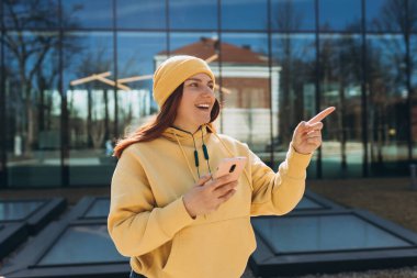 Telefonla şehir geçmişini gösteren mutlu kızıl saçlı kadın. Şehir yaşam tarzı konsepti. Şehir caddesinde yürüyen neşeli genç kadın akıllı telefonu kontrol ediyor..