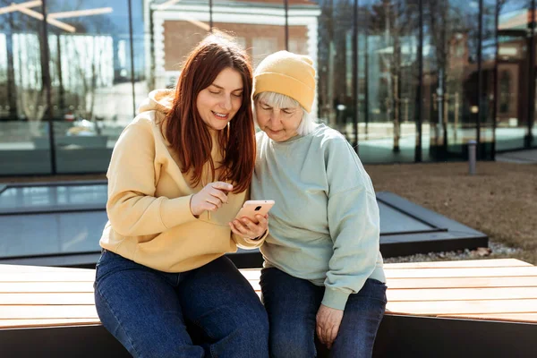 Two women together wear casual clothes using smart phone and sitting on the bench on city street. Young woman and senior middle aged mom spending time together