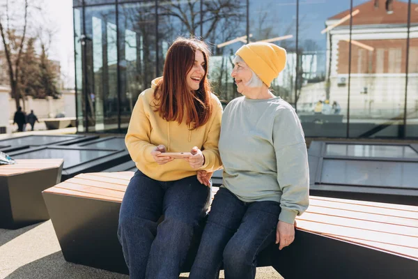 Two women together wear casual clothes using smart phone and sitting on the bench on city street. Young woman and senior middle aged mom spending time together