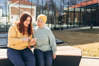 İki kadın birlikte günlük kıyafetler giyer, akıllı telefon kullanır ve şehir caddesinde bankta otururlar. Genç bir kadın ve orta yaşlı bir anne birlikte vakit geçiriyorlar.