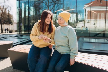 İki kadın birlikte günlük kıyafetler giyer, akıllı telefon kullanır ve şehir caddesinde bankta otururlar. Genç bir kadın ve orta yaşlı bir anne birlikte vakit geçiriyorlar.