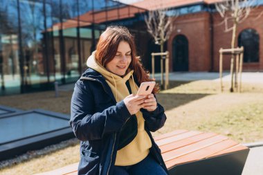 Gündelik giysiler içinde güzel bir kadın akıllı telefonunu şehir caddesinde bankta otururken kullanıyor. Kolej dersinden sonra bankta oturan mutlu ortanca kız..