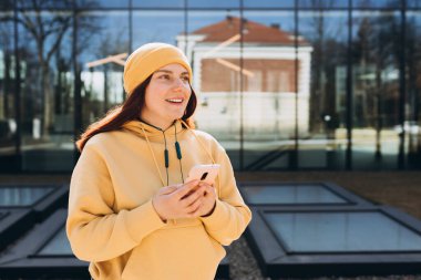 Şehir arka planında akıllı telefonunu kullanan sarı şapkalı güzel bir kadın. Üniversiteden sonra dinlenen mutlu genç kız..