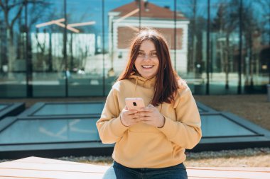 Gündelik giysiler içinde güzel bir kadın akıllı telefonunu şehir caddesinde bankta otururken kullanıyor. Kolej dersinden sonra bankta oturan mutlu ortanca kız..