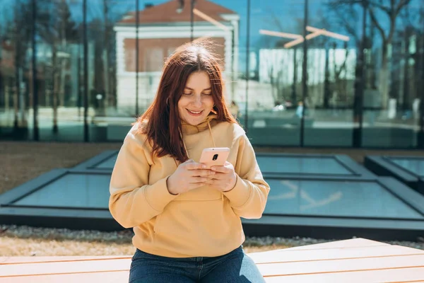 Gündelik giysiler içinde güzel bir kadın akıllı telefonunu şehir caddesinde bankta otururken kullanıyor. Kolej dersinden sonra bankta oturan mutlu ortanca kız..