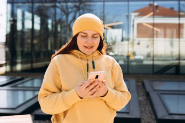 Güzel, kızıl saçlı, mutlu bir gülümsemesi olan bir kadın elinde cep telefonu olan bir şehir sokağında duruyor.