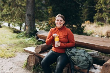 Yürüyüş kıyafetleri içinde kızıl saçlı mutlu bir kadın yürüyüş yaptıktan sonra duraklıyor. Su şişesinden su içen yürüyüşçü ya da termostan sıcak içecek.