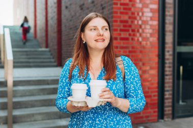 Mavi elbiseli neşeli genç bir kadın dışarıda yürüyor, elinde şehir arka planında kahve fincanı tutuyor. Kentsel yaşam tarzı konsepti.