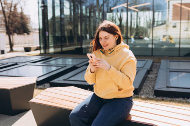 Genç bir kadın sokakta kulaklıkla müzik dinliyor. Kentsel arka planda duran oldukça mutlu kızıl saçlı kadın.