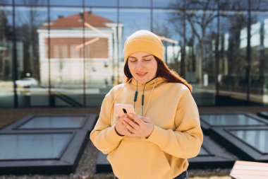 Güzel, kızıl saçlı, mutlu bir gülümsemesi olan bir kadın elinde cep telefonu olan bir şehir sokağında duruyor. Üniversiteden sonra dinlenen genç kız..