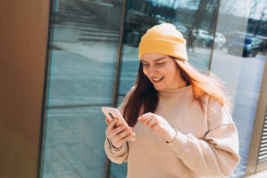 Şehir caddesinde yürüyen neşeli genç kadın akıllı telefonunu kontrol ediyor. Açık havada akıllı telefon kullanan sarı şapkalı güzel bir kadının portresi..