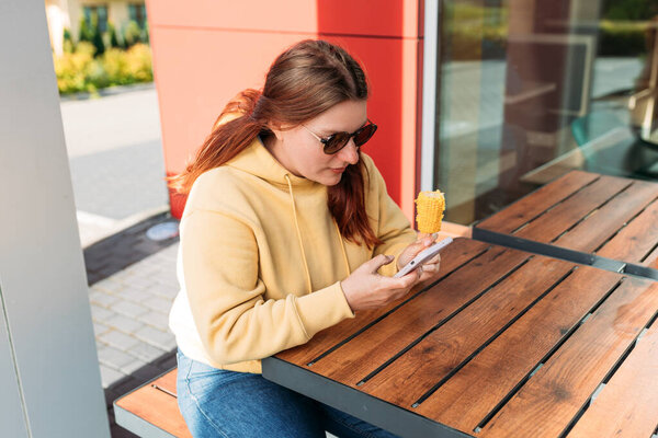 Веселая молодая женщина с телефоном ест кукурузу уличной еды в кафе на открытом воздухе на городском фоне, концепция питания