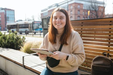 Açık havada tablet kullanan güzel, gülümseyen bir kız öğrenci. Mutlu, genç bir kadın şehir caddesinde bankta oturuyor, online eğitim ya da alışveriş afişi