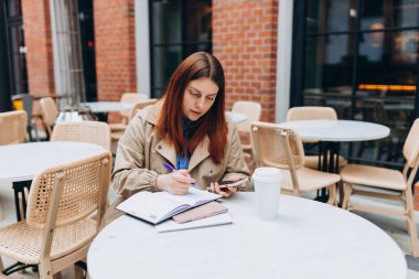 Genç bir kadın kahve içiyor, modern şehir caddesindeki kafe terasında oturuyor. Dışarıda cep telefonu kullanan bir kadın işçi. Masada oturan ve notlar alan biri.